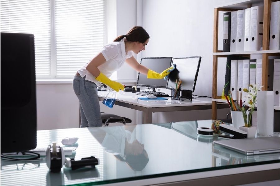 A lady cleaner is wiping the monitor of a computer in an office. In her left hand, she is holding a bottle of cleaning fluid.